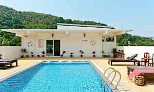 En pool med blått vatten omgiven av solstolar och en vit byggnad i bakgrunden. Grönskande kullar är synliga bakom byggnaden. (AI-genererad text)