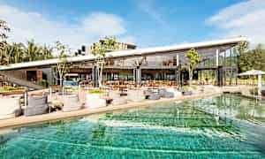 Ett modernt café eller restaurang med stora glasväggar ligger vid en pool under en klarblå himmel. (AI-genererad text)