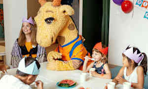 En giraffmaskot sitter vid ett bord med barn som har på sig papperskronor. (AI-genererad text)