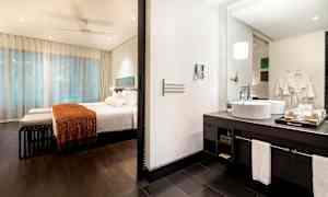 Ett hotellrum med sovdel och badrum. Sovdelen har en säng med vit bäddning och orange filt. Badrummet har tvättställ med spegel. (AI-genererad text)