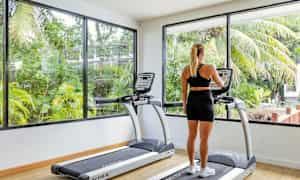 En person tittar på skärmen av en löpmaskin i ett gym med stora fönster och utsikt över grönska. (AI-genererad text)