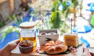 En bricka med olika bakverk och en glasburk med honung, hålls framför en suddig bakgrund av grönska och parasoller. (AI-genererad text)