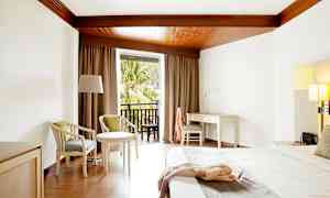 Ett hotellrum med vitt skrivbord, säng och sittmöbler, samt balkong med utsikt över träd. (AI-genererad text)