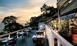 Uteservering med bord och stolar vid kvällstid, med utsikt över träd och hav. (AI-genererad text)