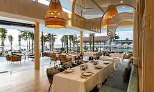 Restaurang med dukade bord och utsikt över palmträd och hav genom stora fönster. (AI-genererad text)