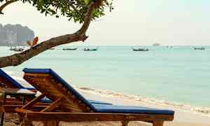 Två solstolar med blå dynor på en sandstrand vid havet, med båtar i bakgrunden. (AI-genererad text)