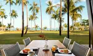 Uteservering med en frukostmåltid vid ett bord. Bakgrunden visar en gräsmatta, palmer och hav. (AI-genererad text)