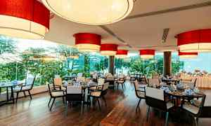 En restaurang med stora röda lampor i taket, fönster med utsikt över grönskande träd och dukade bord. (AI-genererad text)