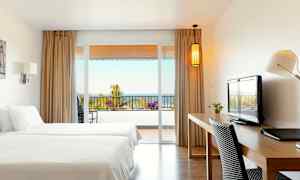 Ett hotellrum med två sängar, skrivbord, och utsikt mot en balkong med hav i bakgrunden. (AI-genererad text)