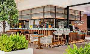 En modern bar med trä- och marmordetaljer, omgiven av gröna växter och sittplatser med stolar och bord. (AI-genererad text)