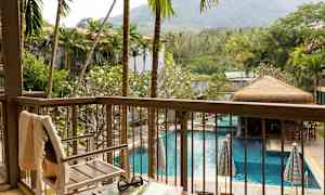 Utsikt från en balkong över en pool omgiven av palmer och gröna växter, med en stol och ett bord i förgrunden. (AI-genererad text)