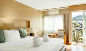 Ett välupplyst hotellrum med en stor säng täckt av vita lakan och kuddar. På sängen finns två handdukar vikta till formen av svanar. Rummet har en träinredning, en sittgrupp med kuddar och en balkong med utsikt mot grönska och byggnader. (AI-genererad text)