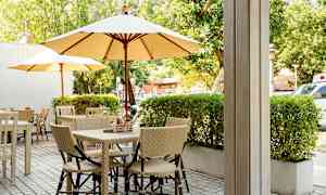 Uteservering med bord och stolar samt parasoll, omgiven av grönska. (AI-genererad text)