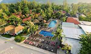 Flygvy av en resort med röda hustak, två poolområden och solstolar nära en sandstrand. (AI-genererad text)