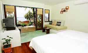 Ett ljust hotellrum med säng, soffa och en stor glasdörr som leder till en terrass med hängmatta och utsikt över havet. (AI-genererad text)