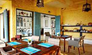 Ett rum med gula väggar och turkosa dörrar, inrett med träbord och rottingstolar. Hyllor med flaskor och serveringsartiklar är synliga. (AI-genererad text)