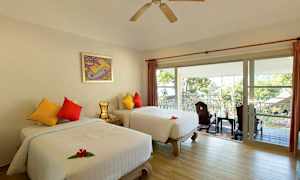 Ett hotellrum med två separata sängar, färgglada kuddar och en balkong med utsikt över träd och ett tak utanför. (AI-genererad text)