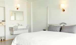 Ett ljust hotellrum med en säng, gråa kuddar, och ett handfat med en spegel. (AI-genererad text)