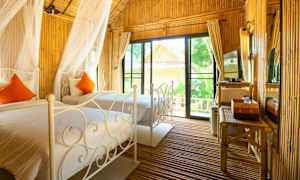 Ett mysigt rum med två sängar, vita metallramar och orangea kuddar. Bambu inredning och stora glasdörrar. (AI-genererad text)