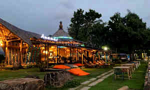 En öppen byggnad med belysning och sittplatser utomhus, omgiven av gräsmatta och träd. (AI-genererad text)