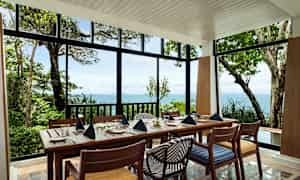 Ett dukat matbord med stolar i en ljus paviljong med utsikt över havet och grönskande natur. (AI-genererad text)