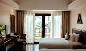 Ett hotellrum med två sängar, ett skrivbord med stol, och stora fönster med utsikt mot en balkong och träd utomhus. (AI-genererad text)