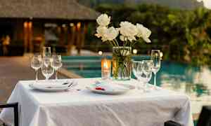 Ett bord dukat med en vit duk, tallrikar, glas och vita rosor i en vas, med en bakgrund av ett poolområde och lummig grönska. (AI-genererad text)