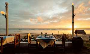 Utsikt över en strand vid solnedgången, med ett dukat bord och stolar i förgrunden. (AI-genererad text)