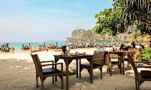 Utomhusservering med bord och stolar vid en strand, med longtailbåtar i bakgrunden och klippor längs kusten. (AI-genererad text)