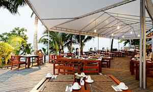 En uteservering med träbord och stolar under ett vitt tygtak, omgiven av tropiska palmer. (AI-genererad text)