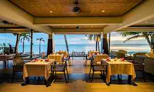 En restaurang vid havet med vita dukar på borden och en utsikt över palmer och stranden. (AI-genererad text)