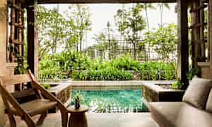 Ett öppet vardagsrum med trästolar och bord, en soffa, och utsikt över en trädgård och en liten pool. (AI-genererad text)