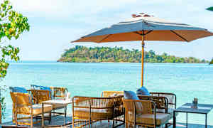 Utemöbler med kuddar under ett parasoll med hav och grön ö i bakgrunden. (AI-genererad text)
