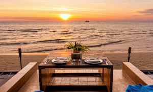 Ett dukat bord med två tallrikar och bestick står vid havet under en solnedgång. (AI-genererad text)