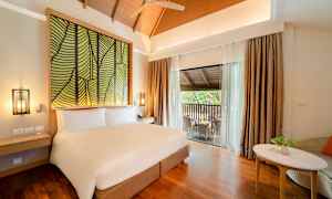 Ett hotellrum med en stor säng, dekorativt huvudgavelmotiv, ett bord med stol samt en balkong med sittmöbler. (AI-genererad text)