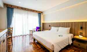 Ett hotellrum med en dubbelsäng, mörka trägolv och ett stort fönster med blå gardiner och vit voil. (AI-genererad text)