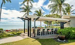 En utomhusbar vid en tropisk strand med palmer och hav i bakgrunden. (AI-genererad text)