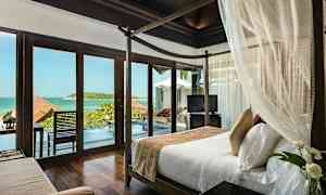 Ett sovrum med en himmelssäng och glasdörrar som öppnar sig mot en utsikt över havet och en veranda med pool. (AI-genererad text)