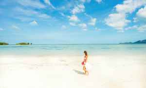 En kvinna går på en ljus sandstrand med havet och en blå himmel i bakgrunden. (AI-genererad text)