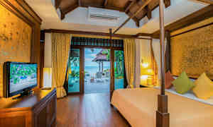 Ett sovrum med trädekoration som har utsikt mot en terrass och havet utanför. Rummet innehåller en säng med färgglada kuddar, ett TV-skåp och lampor. (AI-genererad text)