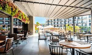 Ett terrassområde med bord och stolar, delvis täckt av tak, med utsikt över en pool och ett hotell. (AI-genererad text)