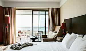 Ett hotellrum med en stor säng, en fåtölj och en balkong med utsikt över havet. (AI-genererad text)