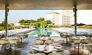 Bild av en utomhusrestaurang med bord och stolar, vid en stor pool omgiven av solstolar och parasoller, med grönska och byggnader i bakgrunden. (AI-genererad text)