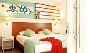 Rum med en dubbelsäng, två gröna kuddar, en röd textil och väggdekoration. (AI-genererad text)