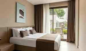 Ett hotellrum med en dubbelsäng, bruna sängkläder och kuddar, samt en glasdörr som leder ut till en terrass med utsikt mot en pool. (AI-genererad text)