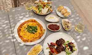 Ett bord med olika maträtter som inkluderar pizza, sallad, bröd, kött med grönsaker, pommes frites samt glas med vitt vin. (AI-genererad text)