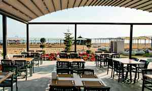 En utomhusservering med bord och stolar, i bakgrunden en sandstrand och havet. (AI-genererad text)