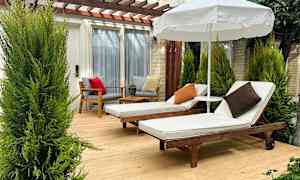 Två solstolar med vita dynor och kuddar står på en träaltan under ett parasoll, omgivna av gröna växter. (AI-genererad text)