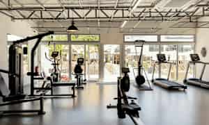 Ett gym med träningsutrustning inklusive löpband, motionscykel och roddmaskin, med glasväggar som visar utsikt över en uteplats. (AI-genererad text)