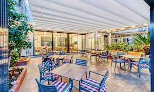 Bild av en utomhusterrass med bord och stolar, samt växter och en inglasad inomhusyta i bakgrunden. (AI-genererad text)